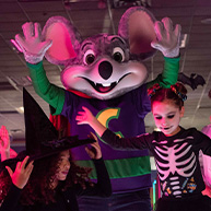 Dos niños disfrazados de Halloween posando con la mascota de Chuck E. Cheese. Una niña disfrazada de bruja de Halloween y un niño disfrazado de un esqueleto de Halloween.